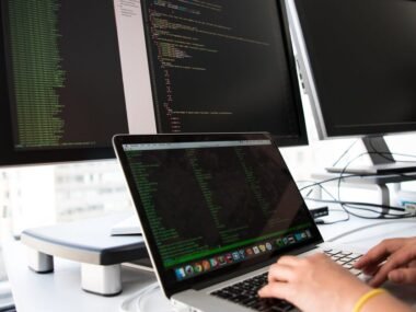 A developer working on a laptop between two monitors showing code